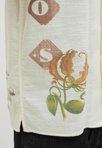 Camisa de manga corta en color crema claro, con estampados florales en marrón y verde, fabricada con un tejido texturizado y con aberturas laterales en el dobladillo.