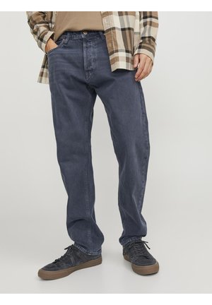 Man draagt donkerblauwe, losvallende jeans, zwarte sneakers, een beige overhemd en een beige-bruine geruite overshirt, staand met handen in de zakken.