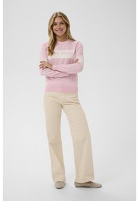 Mujer sonriente con cabello largo y rubio, de brazos cruzados, vestida con un suéter rosa, pantalones de pierna ancha beige y zapatos planos beige con tachuelas.