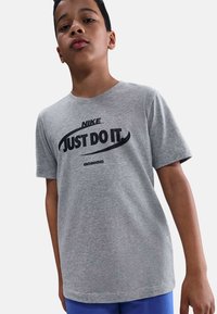 Camiseta de algodón gris con un logo negro de "Nike" y el texto "JUST DO IT" en fuente negrita en el pecho. Mangas cortas, corte relajado.