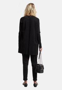 Cardigan lungo nero con taglio dritto, dettagli a coste sul retro, abbinato a pantaloni neri a sigaretta e una piccola borsa trapuntata.