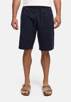 Uomo che indossa pantaloncini blu navy con coulisse e tasche con zip e sandali beige a doppio cinturino, in piedi su fondo bianco.