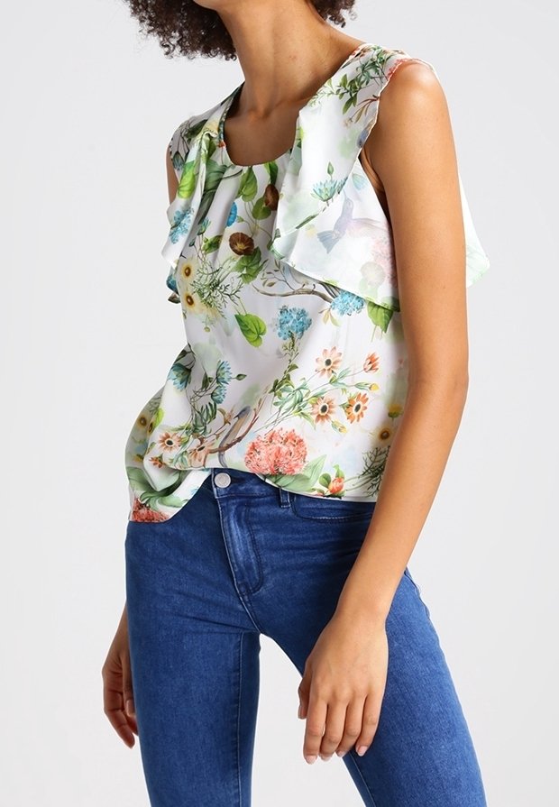 Blouse blanche à fleurs avec col à volants ; présente des imprimés floraux colorés en vert, bleu, orange et rose sur un tissu léger.