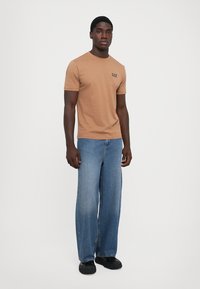 Brun bomull T-skjorte med rund hals og liten EA7-logo, kombinert med lyseblå vide denimjeans og svart fottøy.