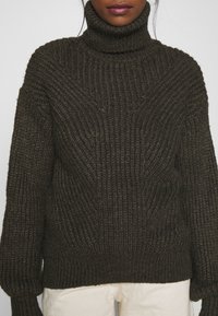 Maglione chunky di un verde scuro con colletto alto, texture a costine e maniche ampie. Presenta una vestibilità rilassata e orlo a costine.