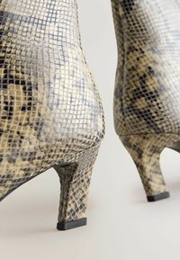 Botines de tobillo con estampado de serpiente en beige y gris con una superficie texturizada. Cuentan con un tacón delgado y un acento negro en la parte inferior de cada tacón.