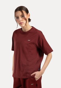 Bordeaux kortärmad t-shirt i bomull med rundad halsringning, sidofickor och en liten varumärkesetikett på vänster bröst.