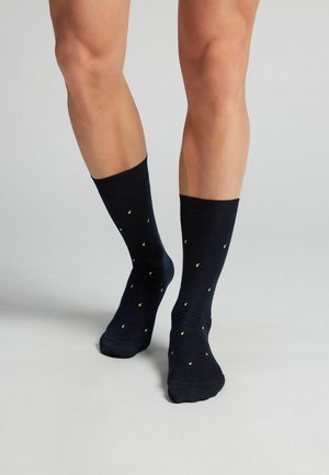 Nohy v černých ponožkách s malými žlutými vzory citronů stojí na světle šedé podlaze před jednobarevným pozadím.