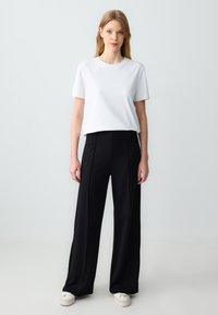 T-shirt blanc à manches courtes associé à un pantalon large taille haute noir. Le modèle porte des baskets blanches ; le tissu semble lisse et décontracté.