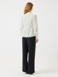 Blusa bianca in pizzo con maniche lunghe, orlo scallop e motivi ad occhiello, abbinata a pantaloni neri a gamba larga, vista da dietro.
