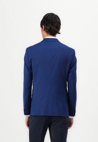 Mørkeblå blazer med en strukturert design, med spiss krage og glatt tekstur. Har to bakventiler og knappede mansjetter.