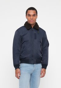 Veste de pilote de marine avec un col en fourrure brun, fermeture éclair, deux poches latérales et un patch sur la poitrine gauche. Tissu lisse, poignets élastiques.