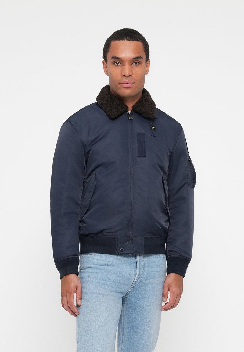Veste de pilote de marine avec un col en fourrure brun, fermeture éclair, deux poches latérales et un patch sur la poitrine gauche. Tissu lisse, poignets élastiques.