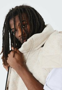 Gequilte vest in lichtbeige met een ruitpatroon. Het heeft een hoge kraag, een ritssluiting en een gestructureerde stof, gedragen over een wit shirt.