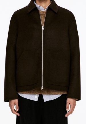 Homme portant une veste zippée marron foncé par-dessus un pull en maille marron et une chemise bleu clair à col, debout avec les bras détendus le long du corps.