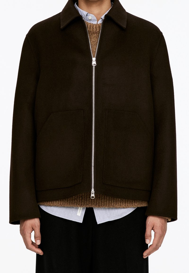 Homme portant une veste zippée marron foncé par-dessus un pull en maille marron et une chemise bleu clair à col, debout avec les bras détendus le long du corps.