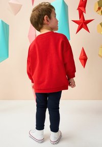 Czerwona bluza z luźnym krojem, granatowe legginsy i białe sneakersy. Tło przedstawia kolorowe geometrie w jasnych odcieniach.