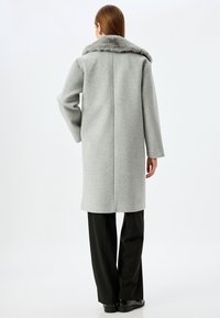 Abrigo de lana gris con collar de piel sintética, de corte recto, mangas largas y costura central en la parte posterior, combinado con pantalones negros.