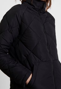 Černá péřová bunda s vysokým límcem, prošívaným vzorem ve tvaru diamantu a zipovým zapínáním. Obsahuje boční kapsy a hladkou, strukturovanou tkaninu.