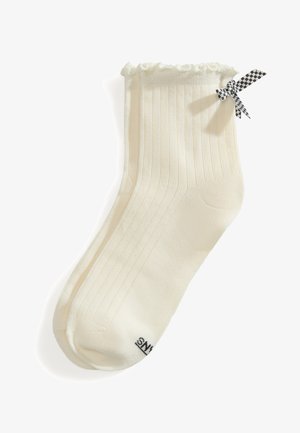 Paar cremefarbene gerippte Knöchelsocken mit Rüschen am Rand und einer schwarz-weiß karierten Schleife auf einer Seite.