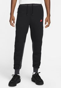 Schwarze Jogginghosen mit seitlichen grauen Paneelen, aus weichem Stoff, mit vorderen Reißverschlusstaschen und einem roten Nike-Logo auf dem linken Oberschenkel.