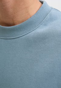 Közeli kép egy világoskék környakú pulóvert viselő személyről, a nyak- és vállrész látható.