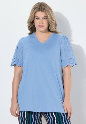 Kvinde med skulderlangt blondt hår iført en lyseblå V-hals top med blonder på ærmerne og stripede bukser mod en ensfarvet hvid baggrund.