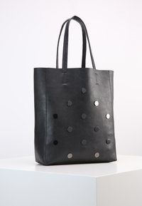 Černá kožená taška přes rameno s plochými stranami, dvěma dlouhými madly a lesklými kulatými kovovými nýty v tečkovaném vzoru na předním povrchu.