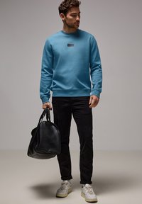 Sudadera azul claro con cuello redondo, tejido texturizado, que presenta una pequeña etiqueta en el pecho. Pantalones negros, bolsa negra y zapatillas blancas.