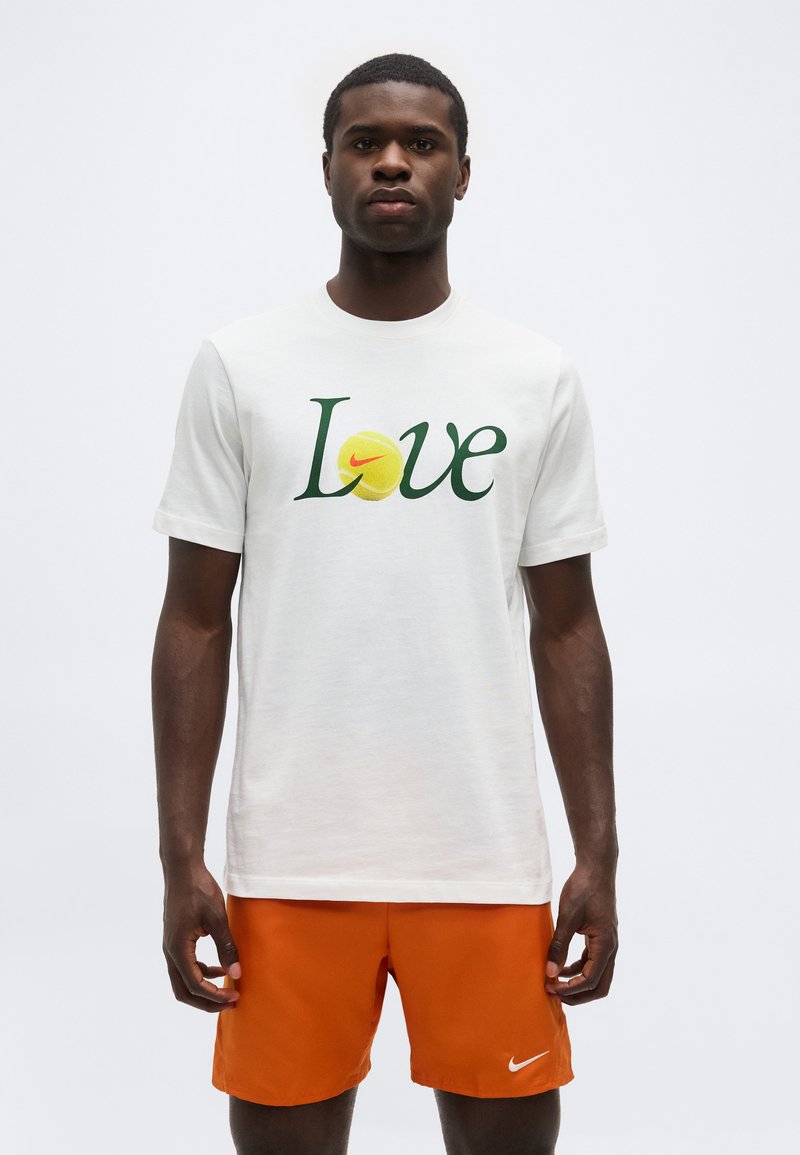 Άνδρας με λευκό μπλουζάκι που γράφει "Love" και το σχέδιο μιας μπάλας τένις, συνδυασμένο με πορτοκαλί σορτς Nike, στέκεται μπροστά σε απλό φόντο.