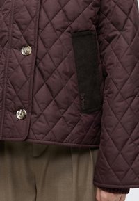 Veste matelassée bordeaux foncé avec couture en forme de losange, deux gros boutons et une poche latérale noire texturée avec le logo Barbour.