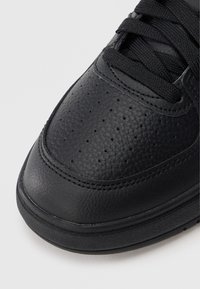 Μαύρο δερμάτινο αθλητικό παπούτσι με υφή στην επιφάνεια, λείο προστατευτικό άκρο  και μαύρα κορδόνια. Διαθέτει τρύπες για διαπνοή και ελαστική σόλα.