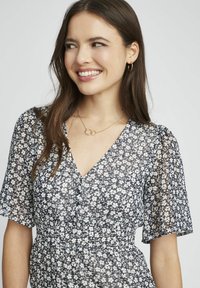 Robe à motif floral avec des manches courtes, décolleté en V et taille froncée. Présente un fond marine avec des fleurs blanches et une texture légère et transparente.
