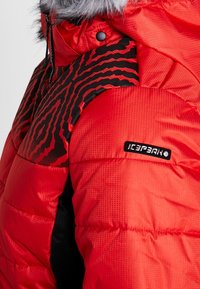 Veste matelassée à motif rouge et noir avec capuche bordée de fourrure et patch logo "ICEPEAK" sur le haut du bras, vue rapprochée de la zone de l'épaule.