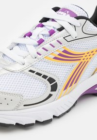 Scarpa da ginnastica bianca con tomaia in rete, accenti viola e gialli, linguetta imbottita, dettagli neri e una suola in gomma testurizzata.