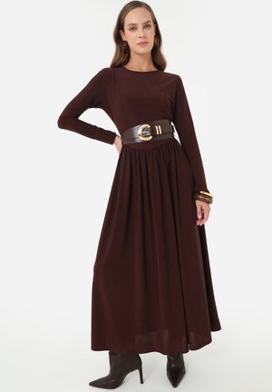 Vrouw draagt een donkerbruine maxi-jurk met lange mouwen, een brede zwarte riem en zwarte, puntige hoge hakkenlaarzen.