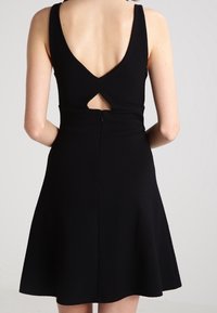 Robe noire sans manches avec un dos en V, dotée d'un détail découpé et d'une jupe évasée. Fabriquée dans un tissu texturé. Fermeture éclair au dos.