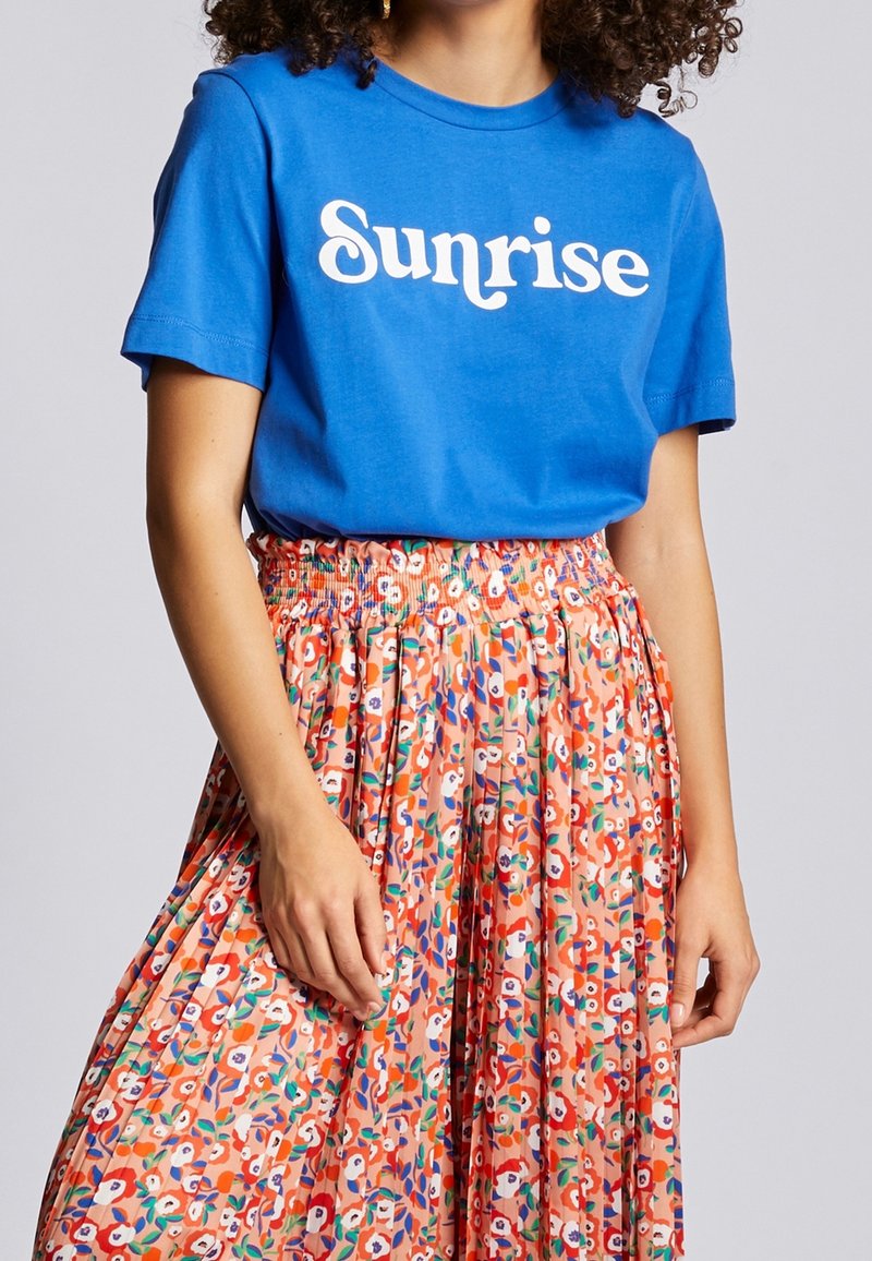 Camiseta de algodón azul con "Sunrise" impreso en blanco, combinada con una falda de flores con plisados en tonos de naranja y verde.