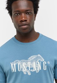T-shirt bleu clair en coton, avec un graphique blanc d'un cheval qui court et le logo "MUSTANG" en lettres majuscules en dessous.