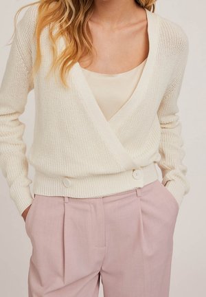 Kobieta ubrana w kremowy dziany sweter z zapięciem na guziki, noszony na beżowej bluzce, oraz jasnoróżowe, wysokowajne, dopasowane spodnie z rękami w kieszeniach.