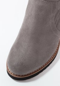 Close-up van een grijze suède bootsneus met een bruin gestikte rand en een zwarte zool op een witte achtergrond.