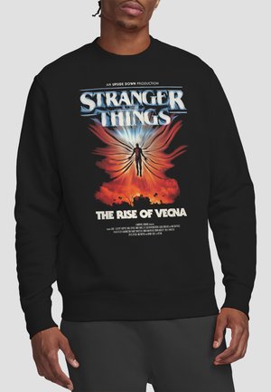 Person wearing a black Stranger Things sweatshirt featuring a glowing figure with wings above a fiery landscape and the text "The Rise of Vecna."