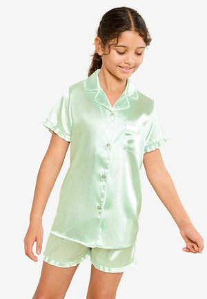 Kurde juustega tüdruk, kes kannab helerohelisest satiinist lühikeste varrukatega pidžaama komplekti koos nööpide ja volangiliste äärtega, naeratab ja vaatab allapoole.