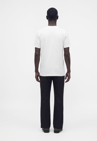 Homme debout, dos tourné, portant une chemise blanche à manches courtes unie et un pantalon noir sur un fond blanc uni.