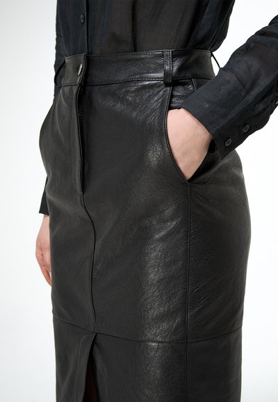 Čierna sukňa z umelej kože s predným rozparkom a pútkami na opasok, kombinovaná s tmavým dlhým rukávom, jedna ruka v vrecku sukne.