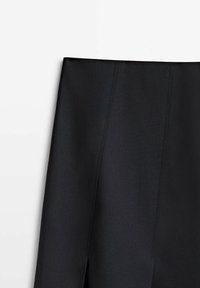 Jupe en tissu noire à taille haute avec une fente à l'avant sur un fond clair uni.