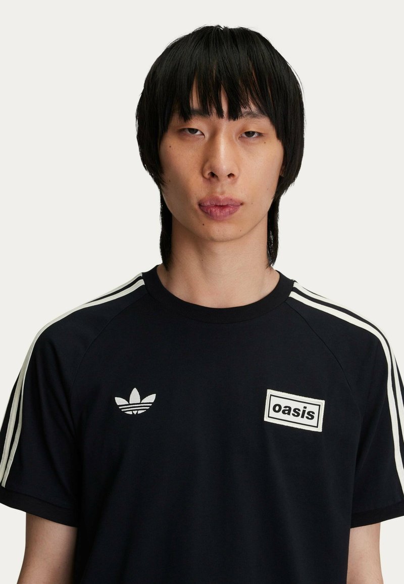T-shirt nera con accenti bianchi, dotata del logo "oasis" sul petto e tre strisce bianche sulle spalle. Realizzata in materiale morbido e leggero.