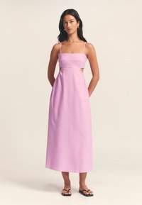 Abito midi rosa in lino con spalline sottili e dettagli a taglio laterale. Presenta un corpetto aderente e una gonna fluida. Sandali piatti indossati.