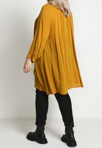 Mosterdgele, losse tuniek met lange mouwen en gathered cuffs, gecombineerd met zwarte slim-fit jeans en zwarte enkellaarsjes.