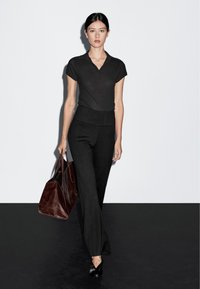 Zwarte getailleerde polo, textuurstof, gecombineerd met hoge zwarte flared broeken. Model draagt een donkerbruin leren totebag.